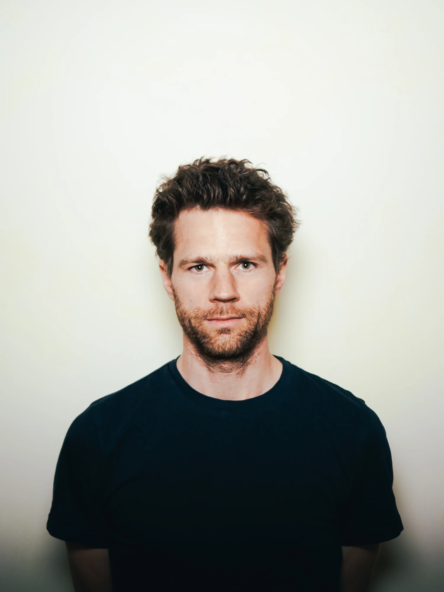 Ein Mann mit dunklem T-Shirt steht vor einem hellen Hintergrund. Er hat kurze, dunkle Haare und einen r&ouml;tlichen Bart.