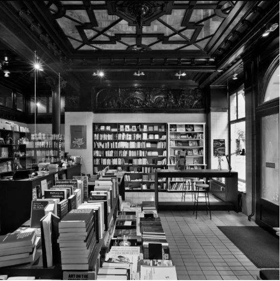 Innenansicht einer Buchhandlung mit vielen Bücherregalen und Büchertischen. Die Decke ist verziert, und es gibt große Fenster.