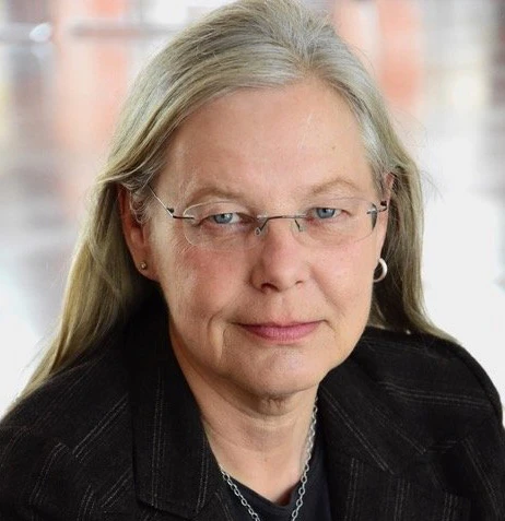 Eine Frau mit Brille schaut in die Kamera. Sie hat lange graue Haare und tr&auml;gt eine schwarze Jacke.