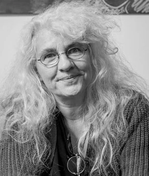 Frau mit Brille und lockigen, langen, grauen Haaren l&auml;chelt. Sie tr&auml;gt eine Halskette und einen gestrickten Pullover. Das Bild ist in Schwarz-Wei&szlig;.