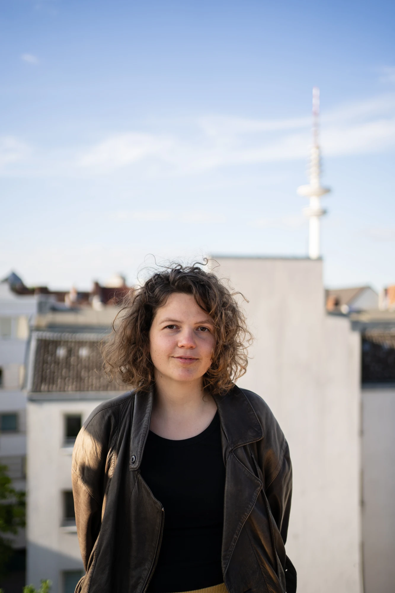 Eine Person mit Locken und kinnlangen Haar steht vorne. Sie tr&auml;gt eine dunkelbraune Jacke und ein schwarzes Oberteil. Im Hintergrund sieht man Himmel und Geb&auml;ude.