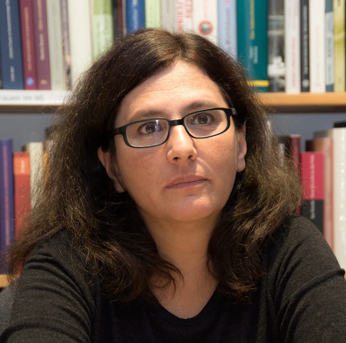 Eine Frau mit Brille schaut nachdenklich. Sie hat dunkle, lockige Haare und tr&auml;gt einen schwarzen Pullover. Im Hintergrund sind B&uuml;cherregale.