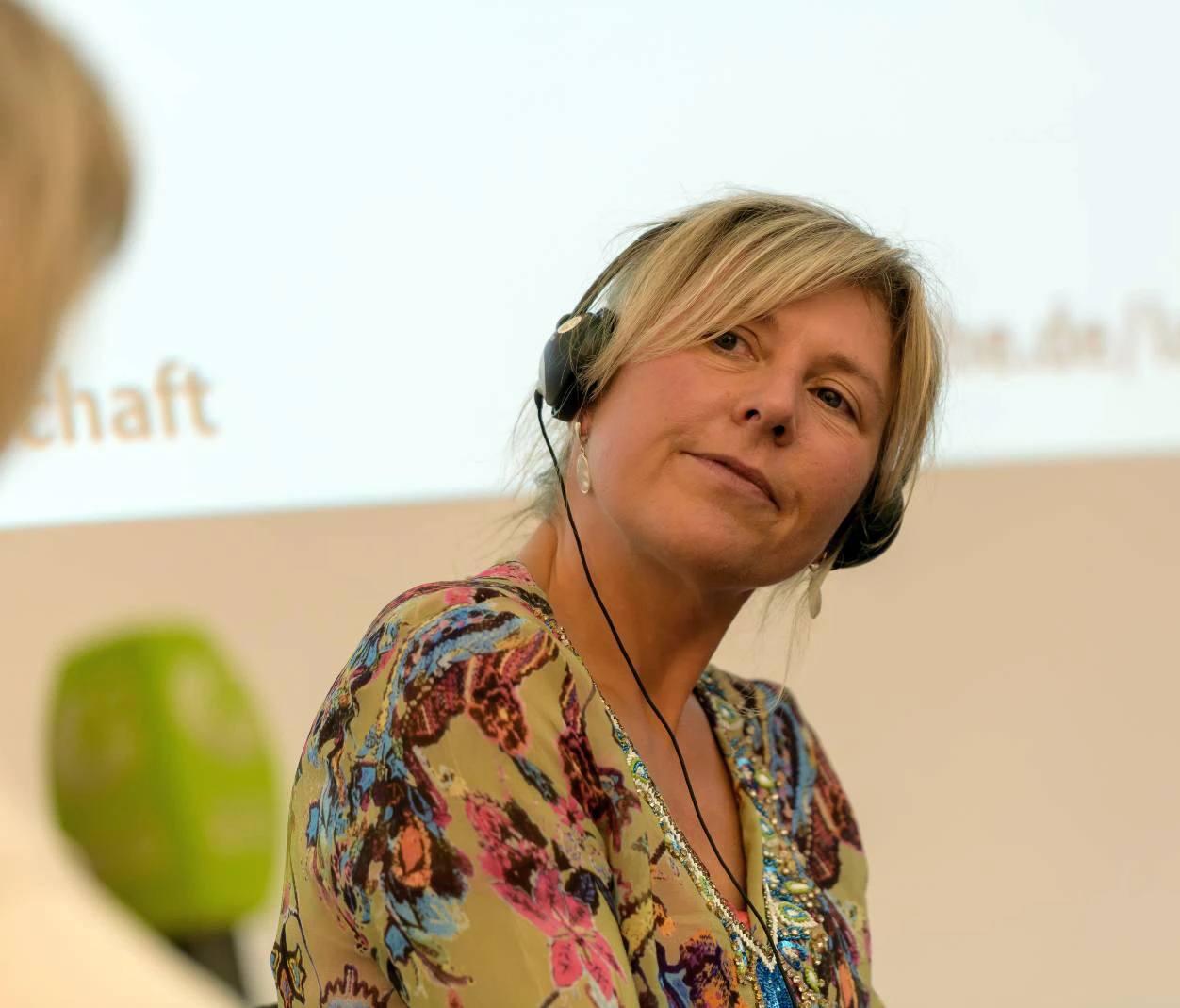 Frau mit Kopfh&ouml;rer bei einer Veranstaltung. Sie tr&auml;gt ein buntes Oberteil und sieht konzentriert aus.