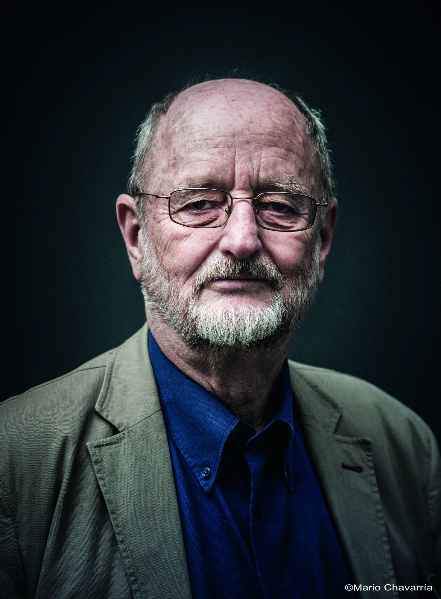 Ein &auml;lterer Herr mit Brille, Glatze und Bart schaut in die Kamera. Er tr&auml;gt ein blaues Hemd und eine beige Jacke. Der Hintergrund ist dunkel.