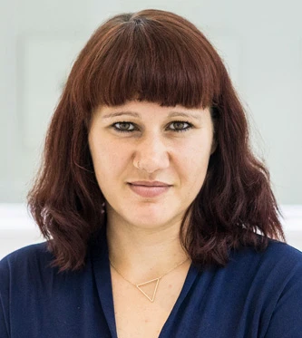 Eine Frau mit schulterlangem braunem Haar und einem blauen Oberteil l&auml;chelt in die Kamera. Sie tr&auml;gt eine Halskette mit einem geometrischen Anh&auml;nger.