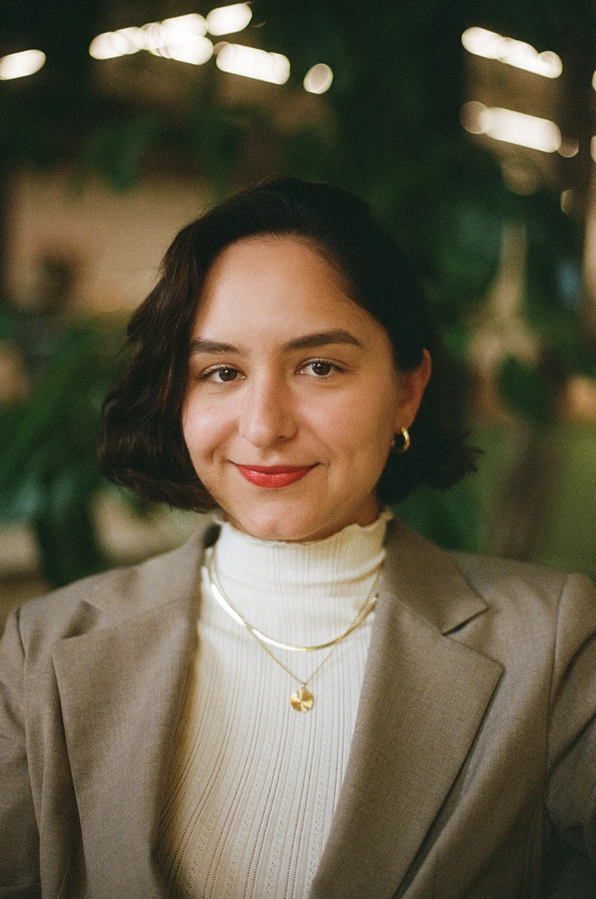 Ein freundlich l&auml;chelnde Person mit kurzen dunklen Haaren. Tr&auml;gt ein wei&szlig;es Rollkragenoberteil, eine helle Jacke und eine goldene Halskette. Der Hintergrund ist unscharf mit Lichtern.
